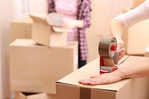 woman sealing a box with tape and a woman holding paper behind her