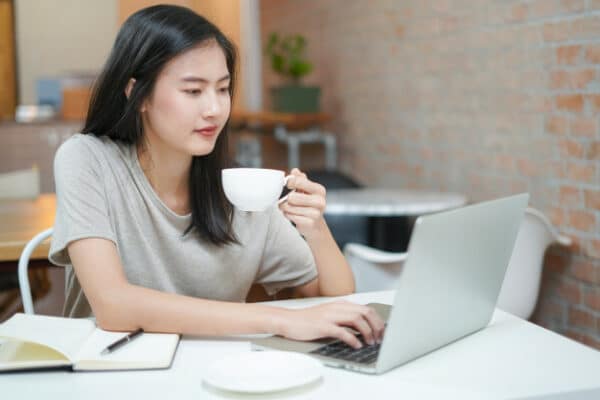 an asian woman drinking tea while working on her laptop